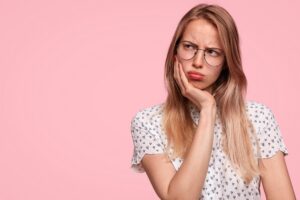 Discontent thoughtful Caucasian female has grumpy expression, keeps hand under chin and looks pensively aside, wears glasses and white blouse, poses against pink background with blank copy space